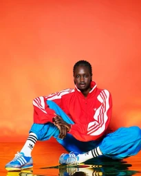 Black woman seated cross-legged on a shiny reflective orange surface wearing a red tracksuit and blue trousers
