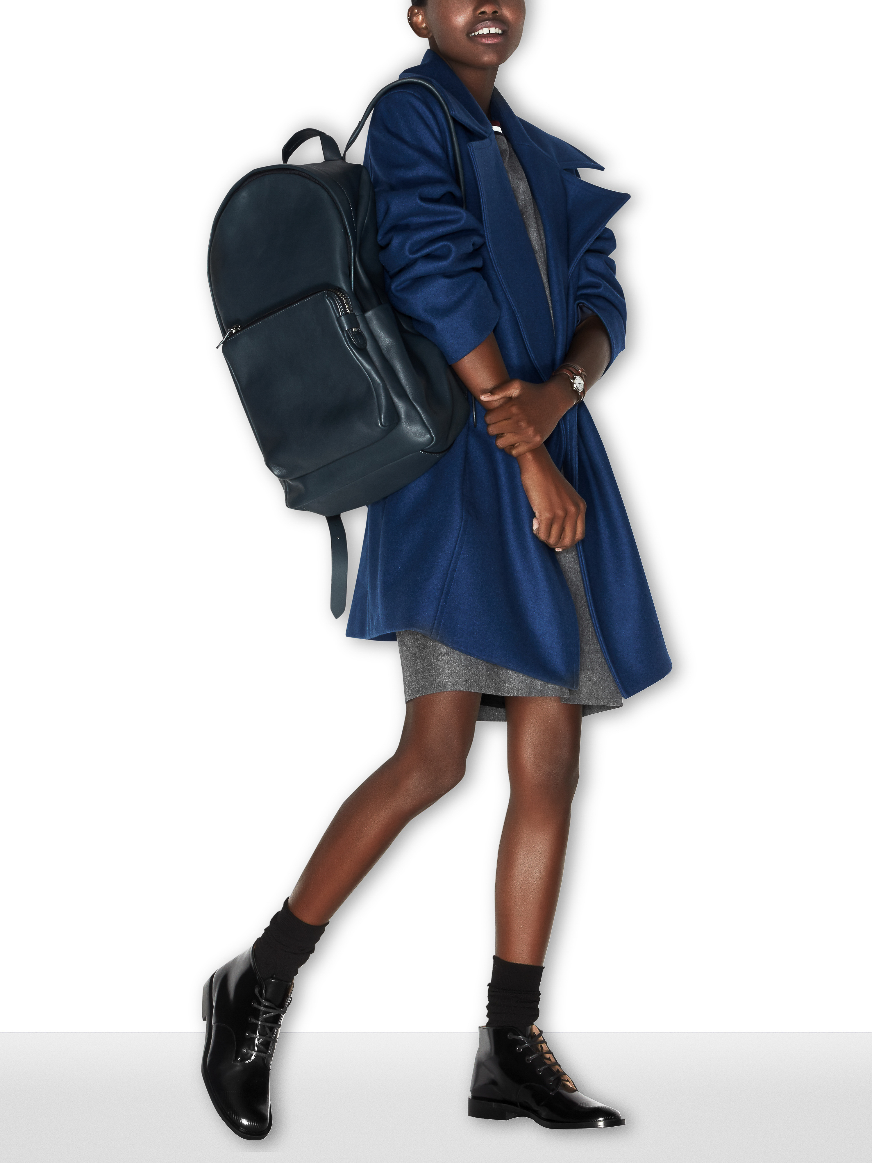 Stylish African woman stands in a blue coat and black boots carrying a black backpack against a stark white backdrop, fashion shoot.