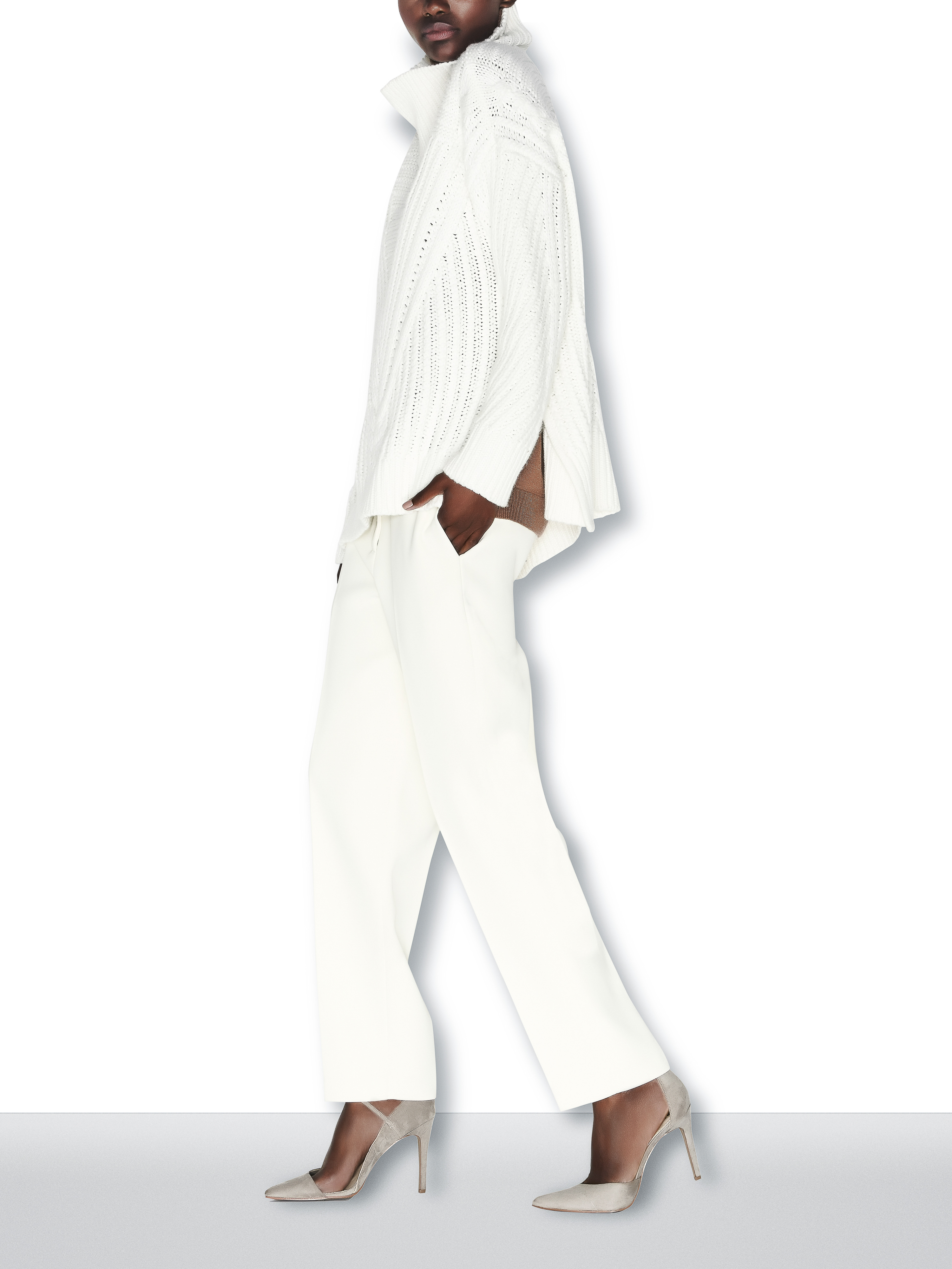 An adult woman is walking in a studio wearing a white sweater and pants with heels. The background is solid white.