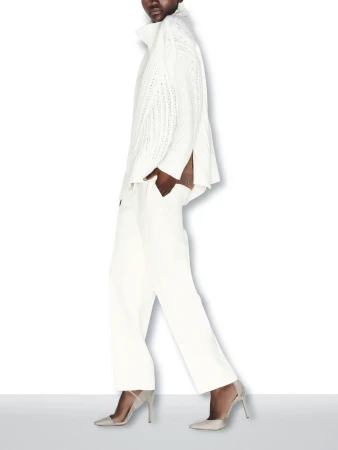 An adult woman is walking in a studio wearing a white sweater and pants with heels. The background is solid white.