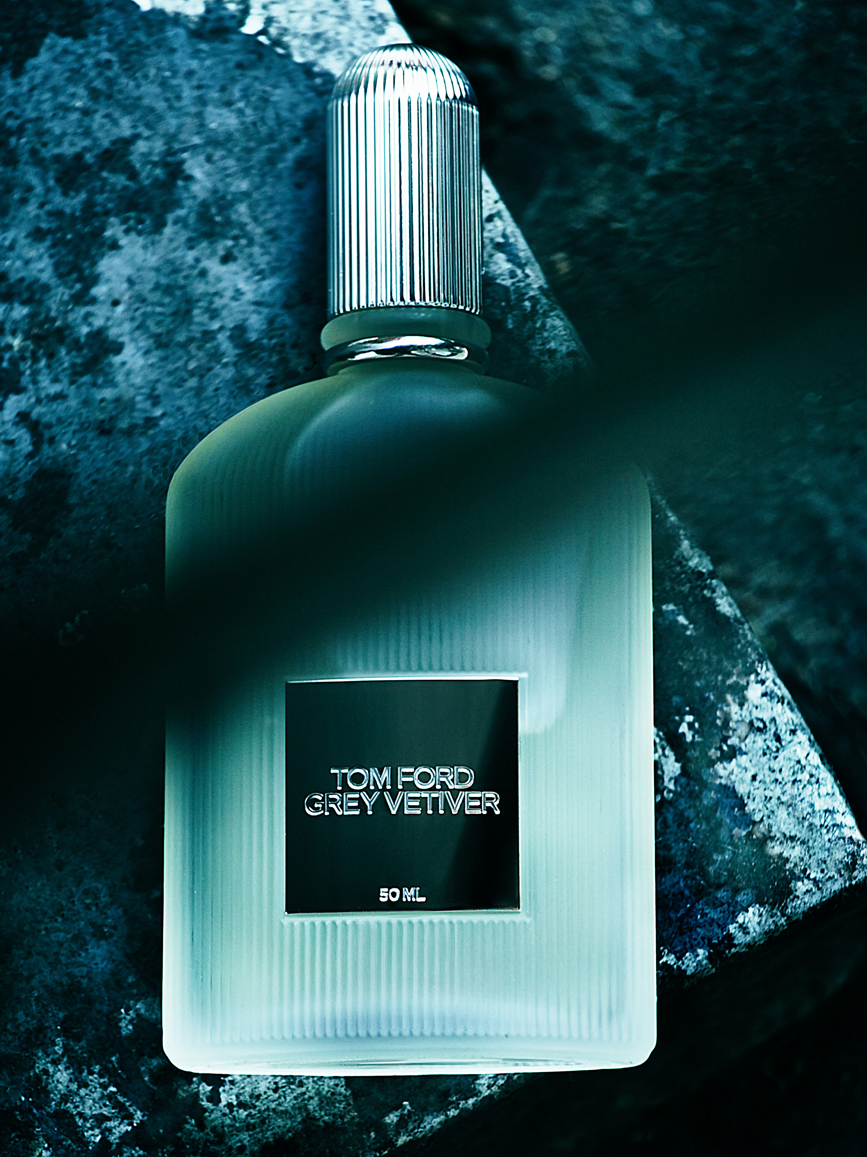 Close-up of a Tom Ford Grey Vetiver fragrance bottle with a ribbed silver cap, set against a textured blue and gray stone surface.