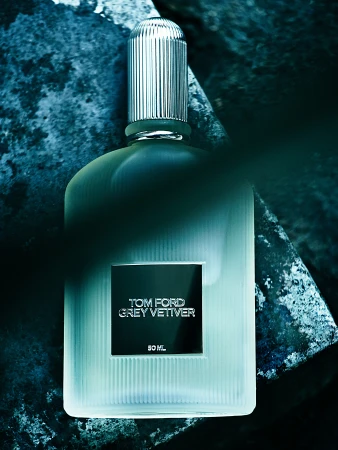 Close-up of a Tom Ford Grey Vetiver fragrance bottle with a ribbed silver cap, set against a textured blue and gray stone surface.
