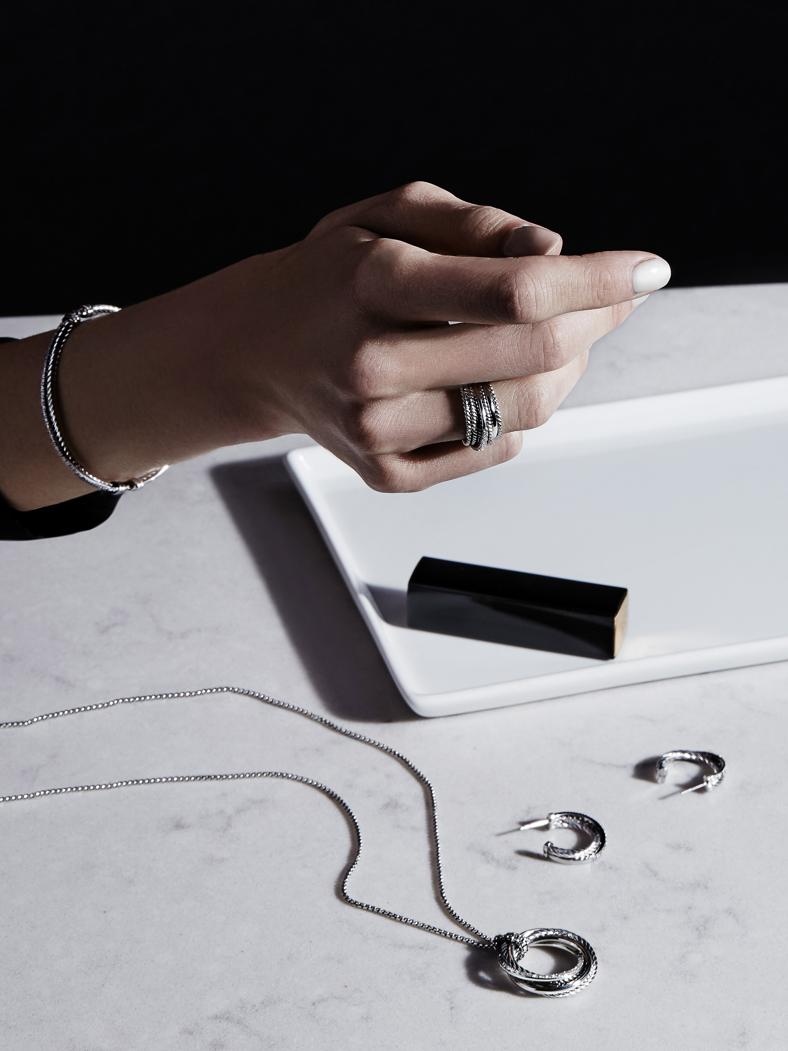 Close-up shot of adult female hands displaying silver jewelry, including rings, a bracelet, a necklace, and hoop earrings on a marble surface.