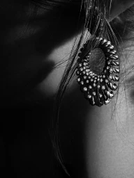 Close-up black and white photograph of an ornate diamond-encrusted hoop earring worn against dark hair and dramatic shadows.