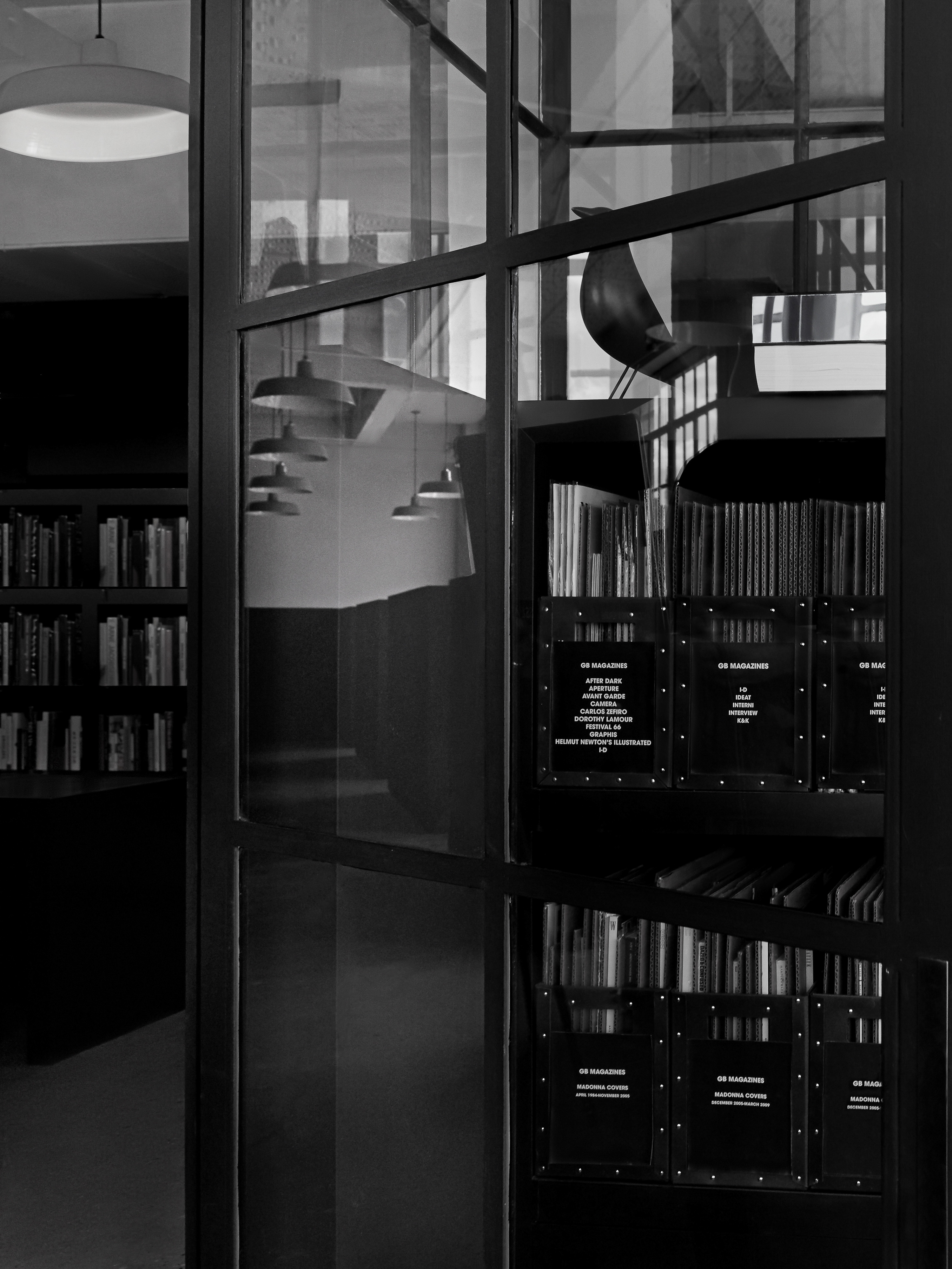 Black and white photo of a modern interior with books on shelves, hanging pendant lights, and reflections in glass panels.