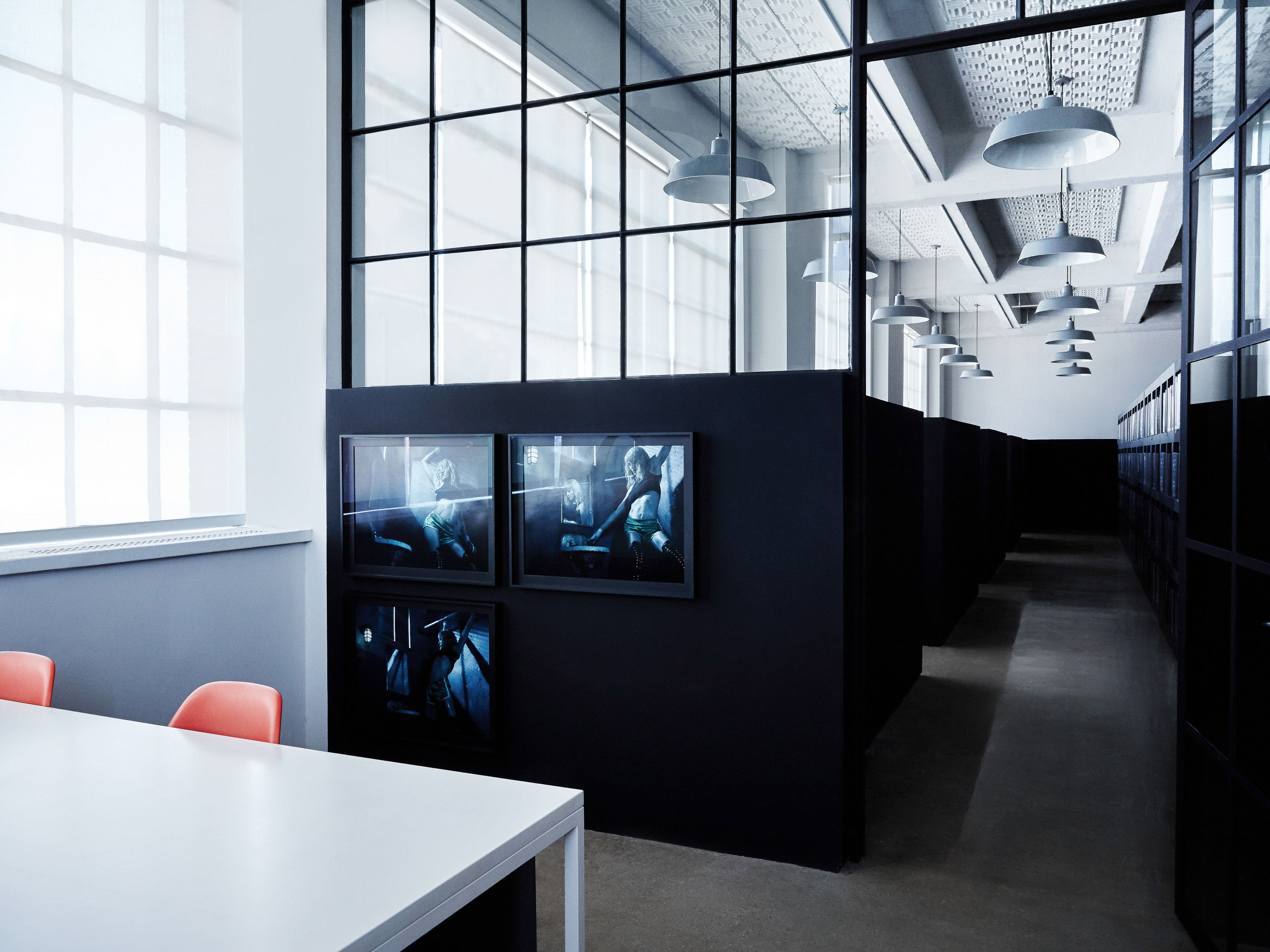 Modern office interior with white table, orange chairs, black room dividers, large windows, and hanging pendant lights.