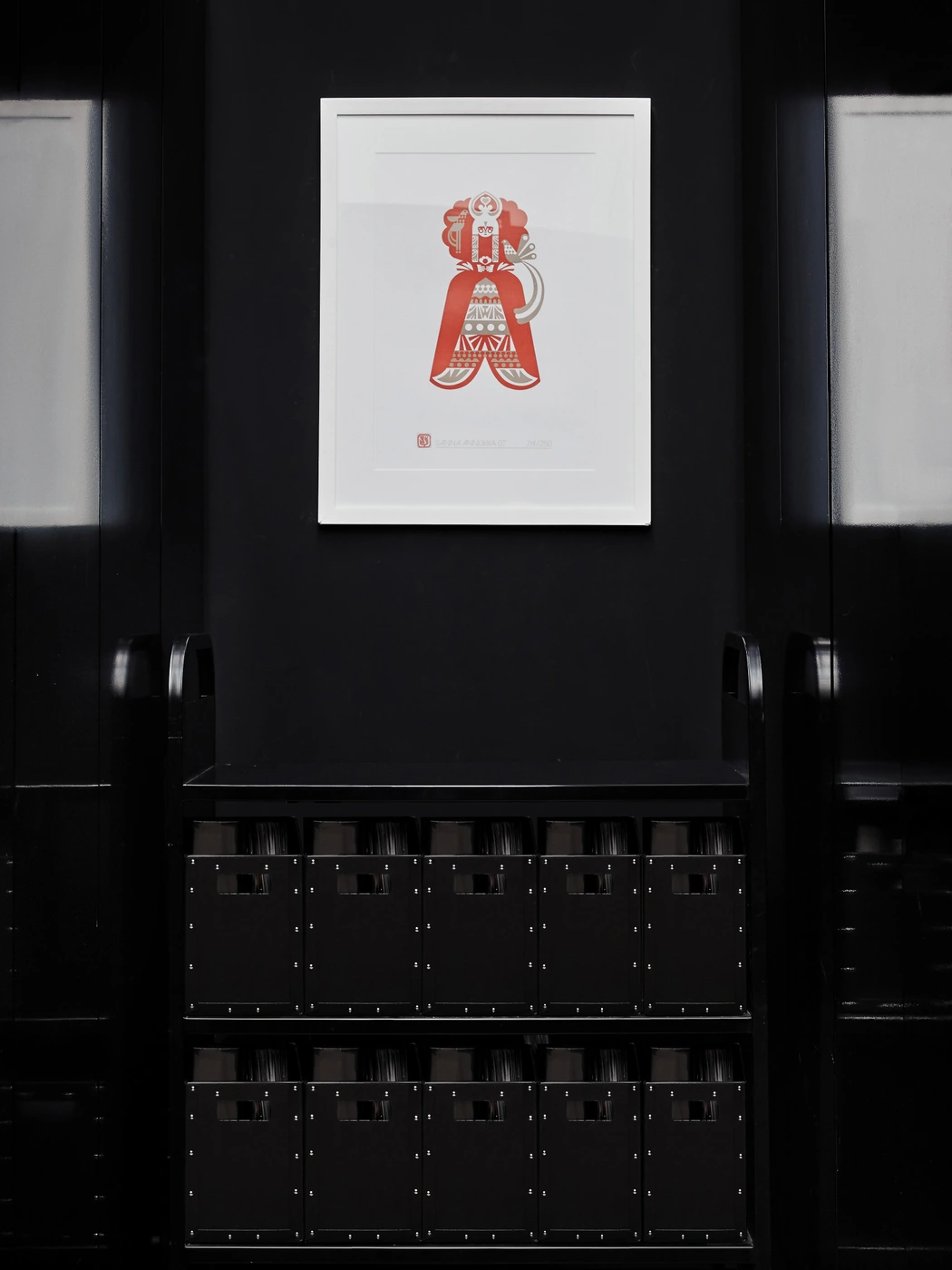 Framed red graphic print in a white frame hanging on a dark wall above a black storage rack filled with organized glassware.