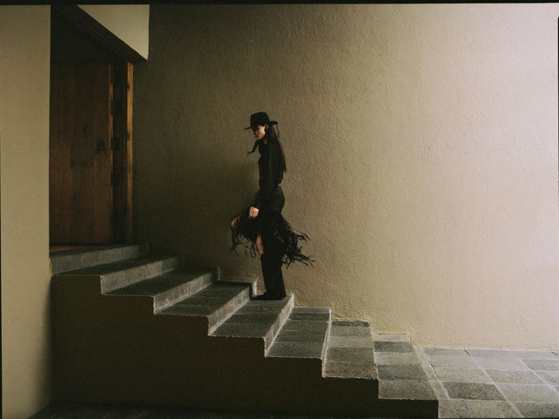 A woman in a black outfit descends stone stairs in a dimly lit outdoor area with a beige wall and a wooden door on the side.