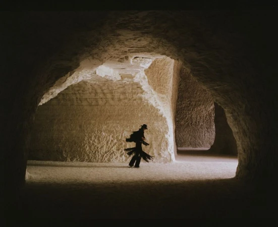 Person in dark clothing stands in a large, dimly lit cave with tan walls and ceiling, creating a mysterious and surreal atmosphere.