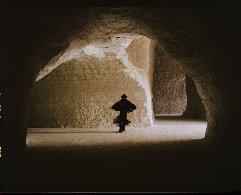 Person in black dress stands in a vast cave with textured walls and archways, creating a dramatic and somewhat mysterious atmosphere.