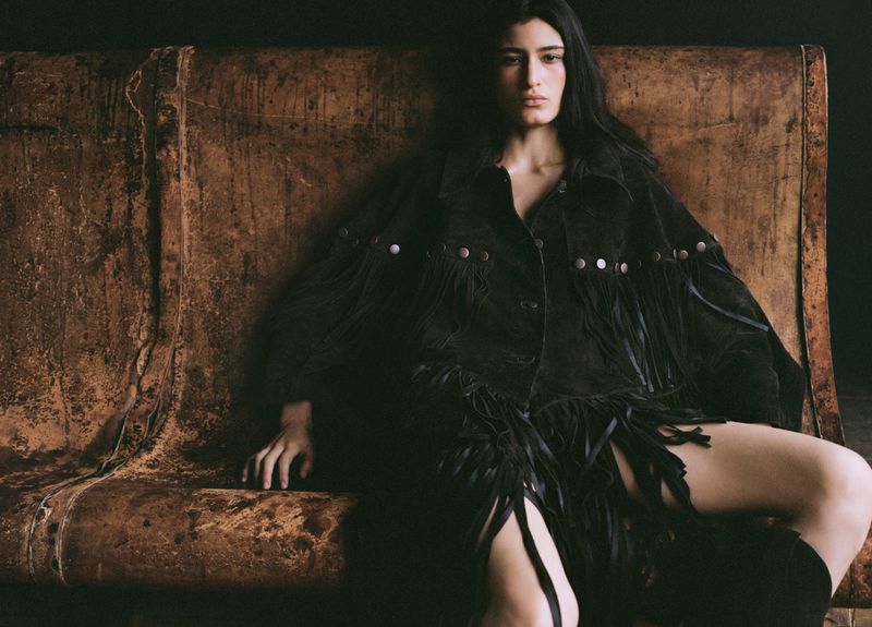 A woman sits on a weathered surface wearing a black fringe jacket and boots in a dimly lit, indoor environment.