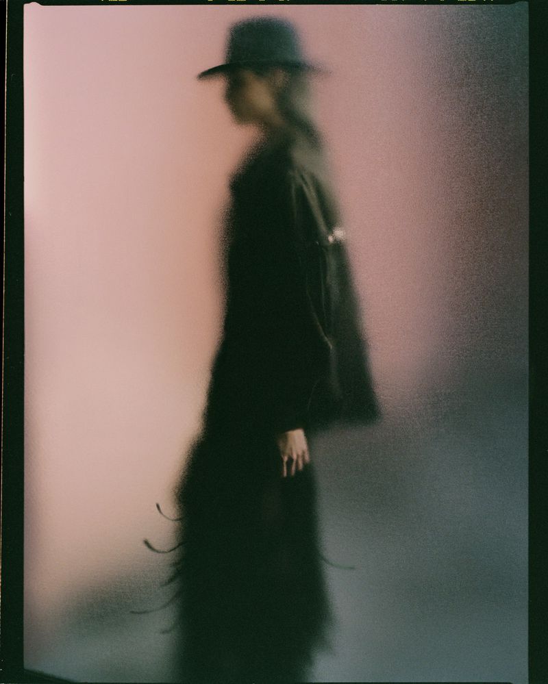 Fashionable woman in hat and long coat poses in a blurred pink-toned environment, artistic fashion editorial, studio setting.