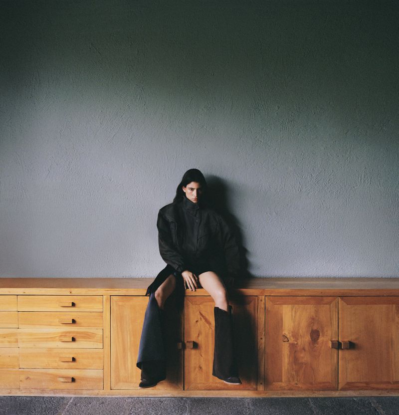 A woman in an oversized black coat and boots sits on a wooden cabinet against a gray wall in a high-fashion, moody editorial shot.