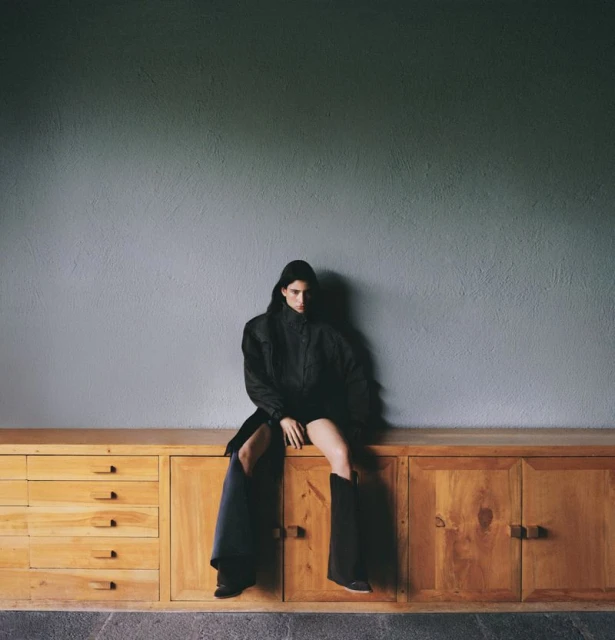 A woman in an oversized black coat and boots sits on a wooden cabinet against a gray wall in a high-fashion, moody editorial shot.