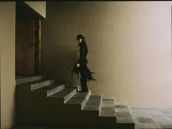 Model Luiza Perote in an all-black outfit with a brimmed hat and fringe accents walking up outdoor stone stairs toward a doorway.