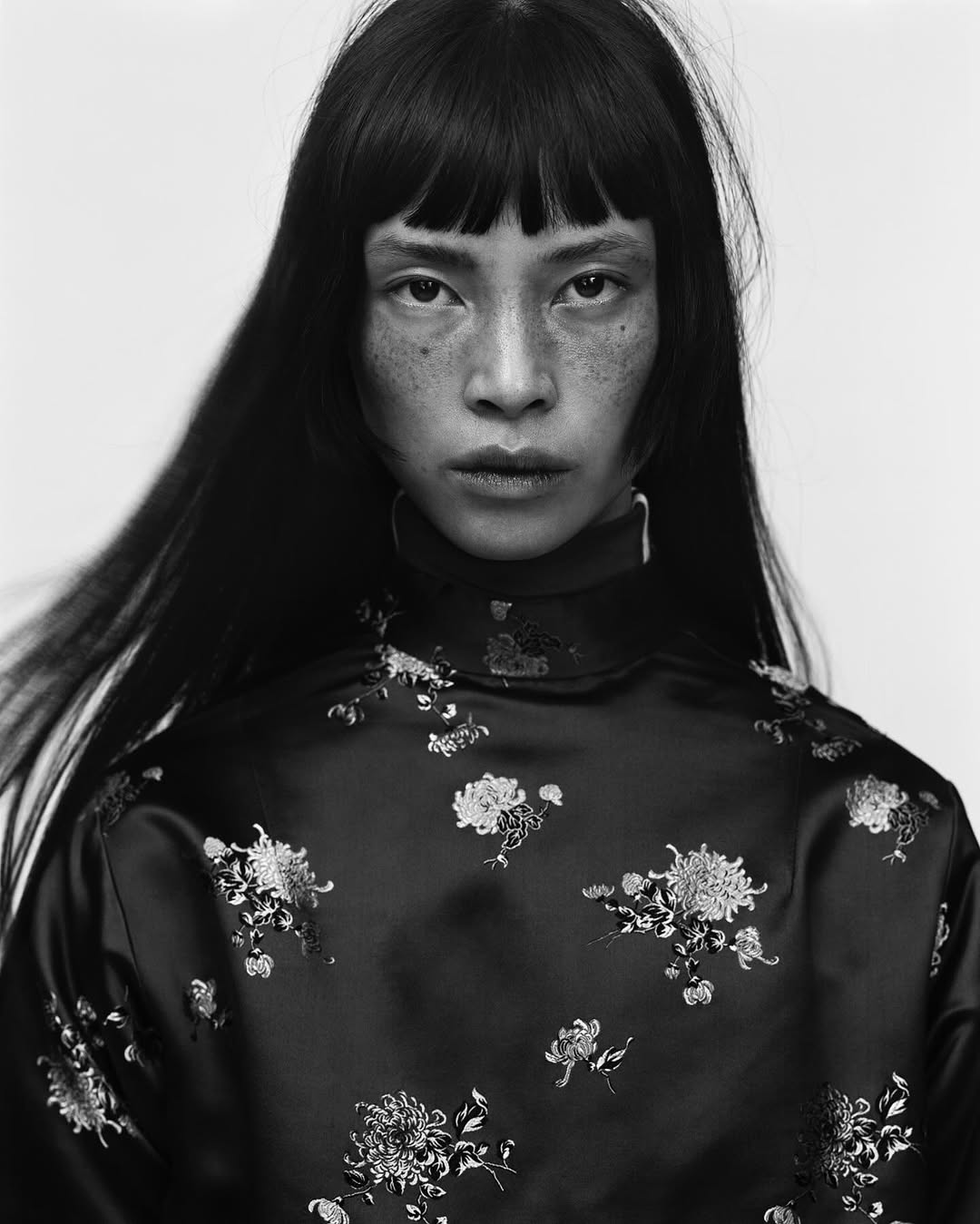 Portrait of an Asian woman with dark hair and freckles wearing a floral-patterned top against a white backdrop in a studio.