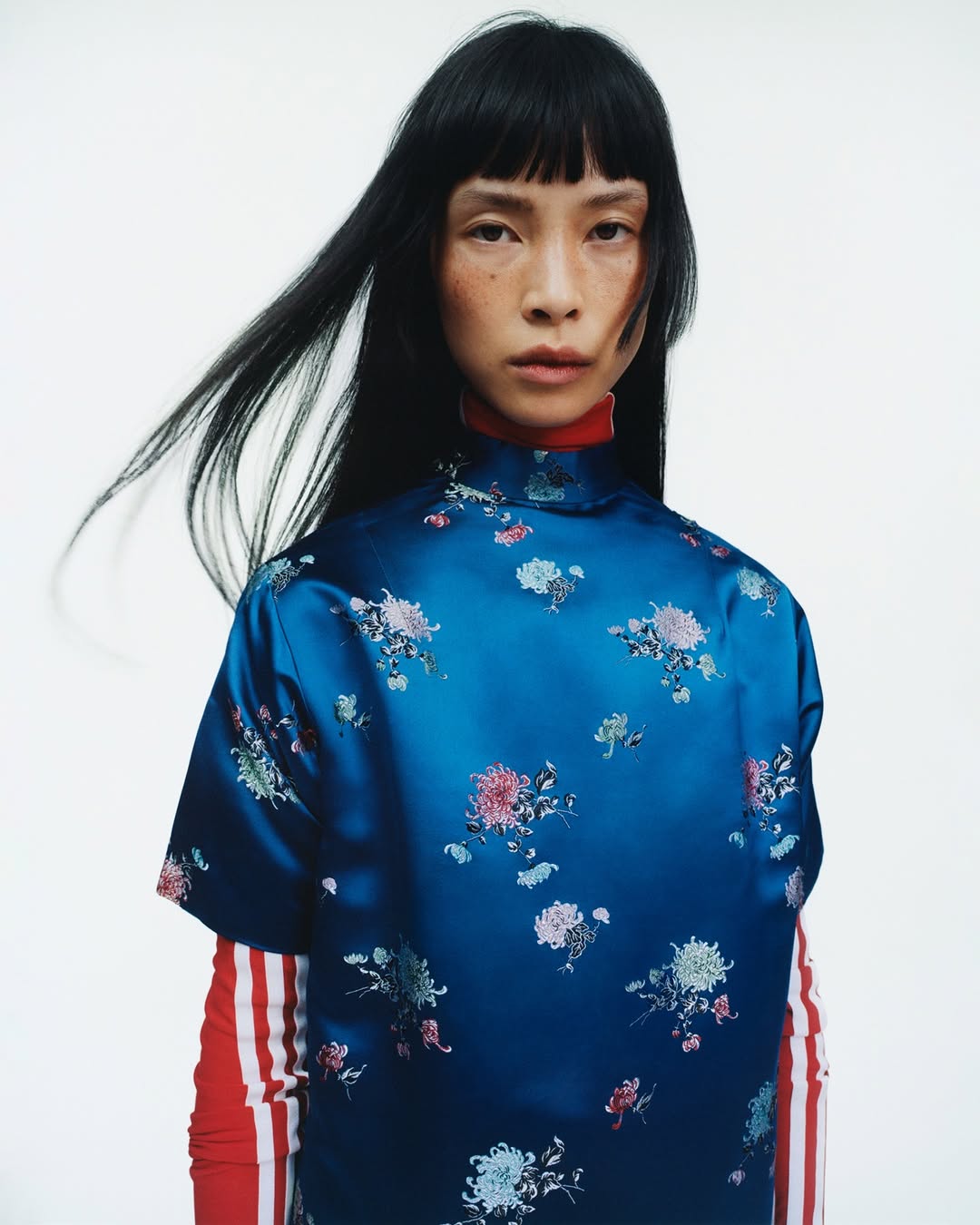 Portrait of an Asian woman wearing a patterned blue satin top with a red mock turtleneck and black hair against a white background.