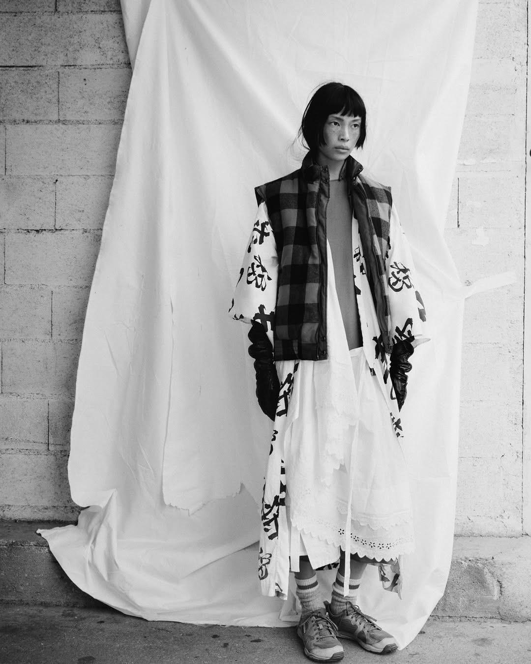 Monochrome fashion shot: An Asian woman poses against a white sheet and brick wall in a layered outfit with a vest and white dress.