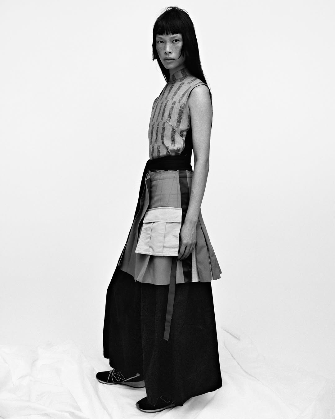 Monochrome portrait of a slender Asian female model in avant-garde fashion, featuring a striped top, tiered skirt, and wide-leg pants, studio lit.