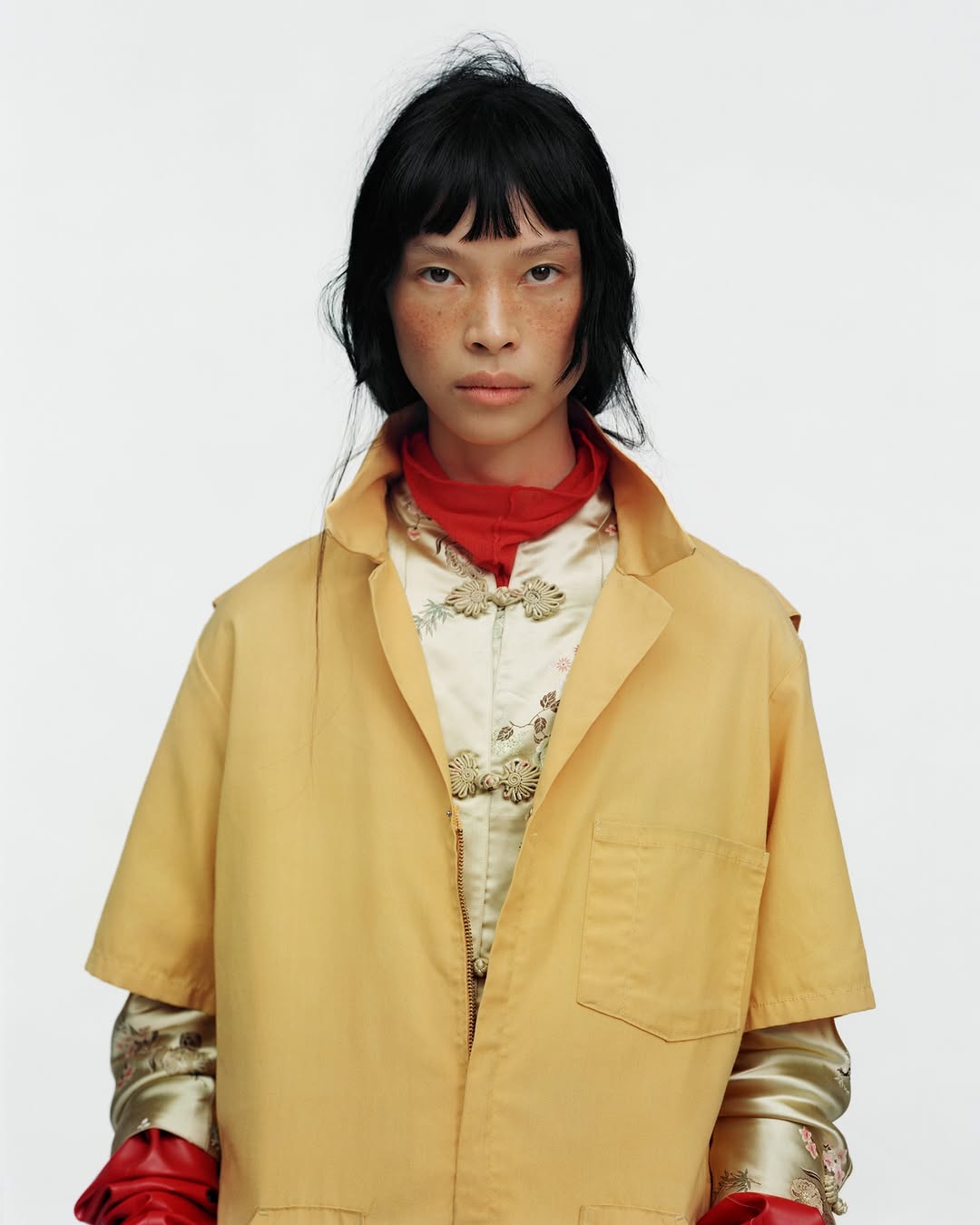 Fashion portrait of an Asian woman with black hair wearing a tan jacket, red scarf, and vintage-style floral top against a white backdrop.