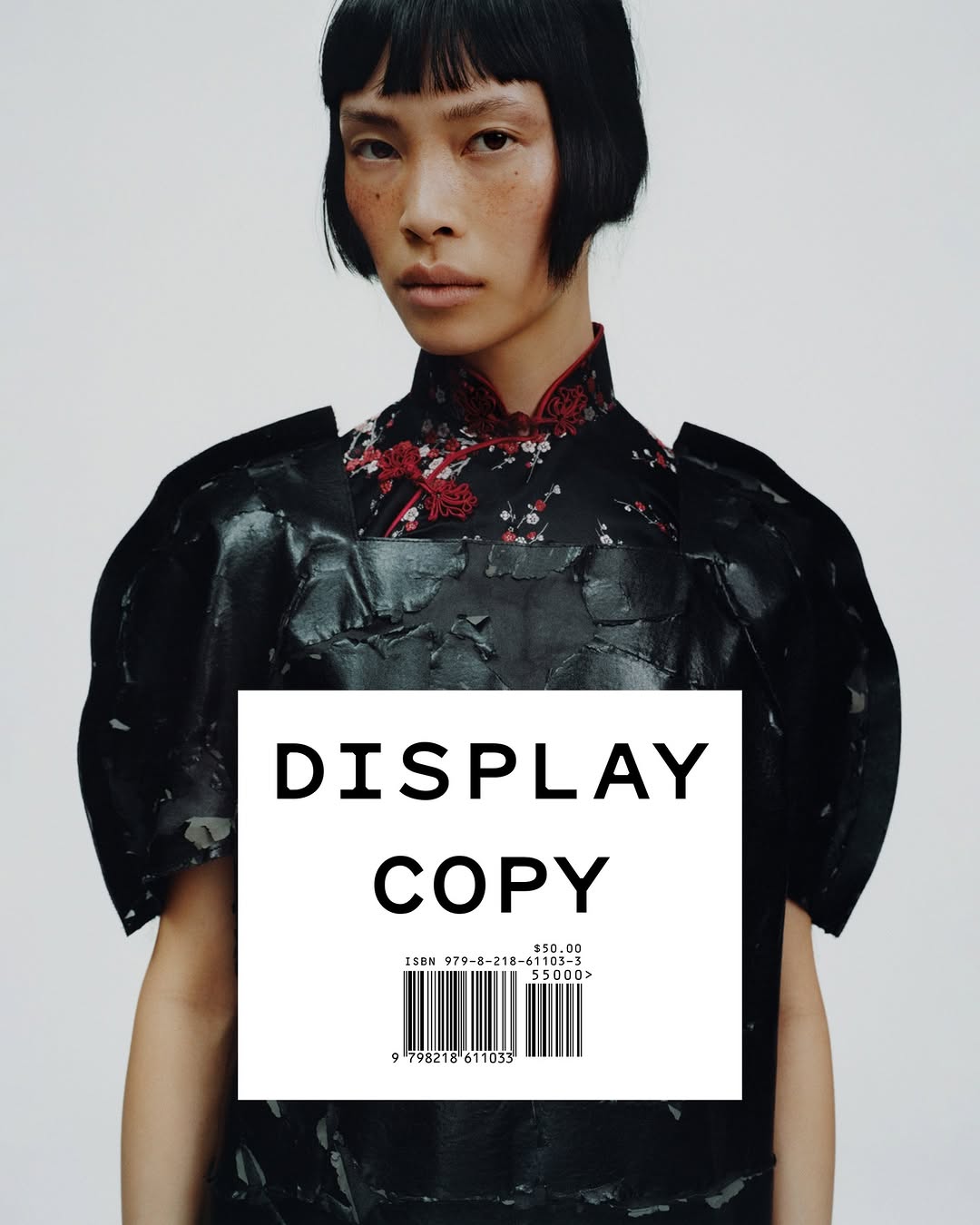 Close-up of a young Asian woman with short black hair, fair skin, wearing a dark, avant-garde top with a floral pattern on a white background.