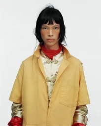 Model with dark hair wearing a yellow coat over patterned top and red gloves against white background