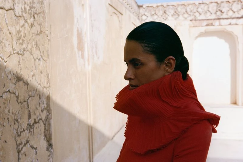 Woman poses in red high-collar top against a textured neutral wall with an arched doorway and bright sky in the background, outdoor setting.