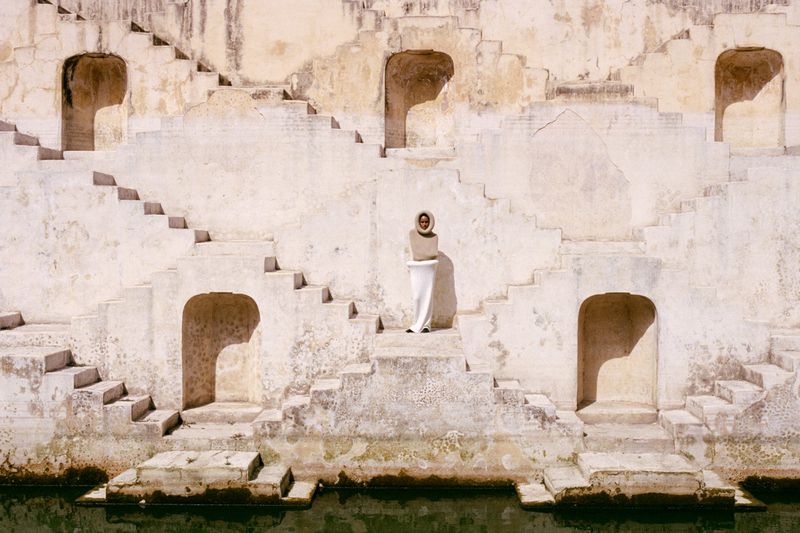Adult female in a white dress with stairs in the background. Ancient architecture with repeating steps leads to a pool of water below.