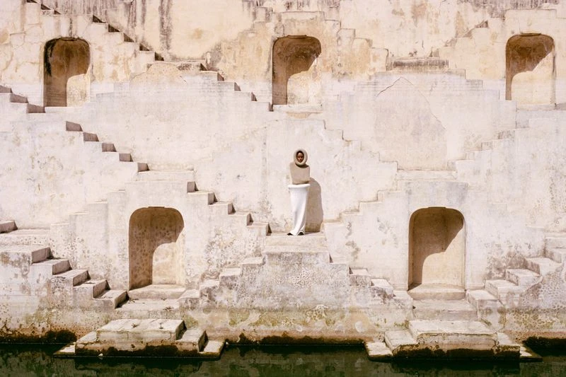 Adult female in a white dress with stairs in the background. Ancient architecture with repeating steps leads to a pool of water below.