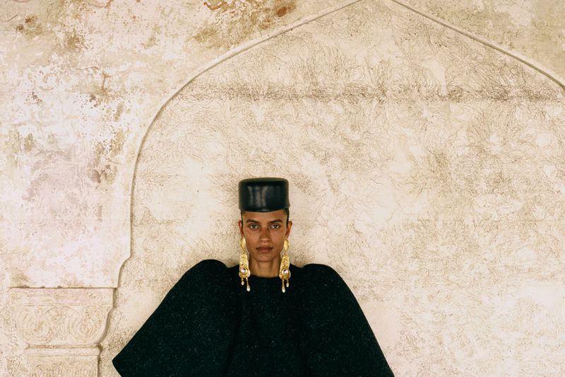 Portrait of an adult female wearing a black cap and large gold earrings, posing in front of an off-white patterned wall with arch.