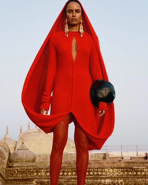 Fashion shot of an adult woman wearing a red hooded dress, fishnet tights, and statement earrings against a sky and distant architecture.