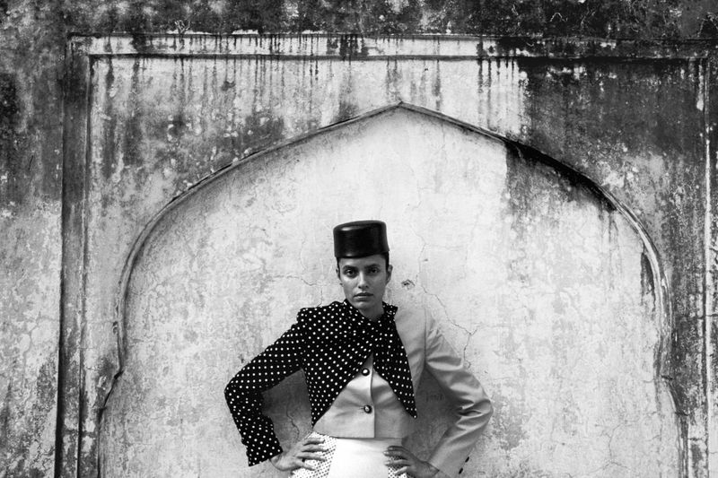 Lakshmi Menon in a polka dot jacket and pillbox hat posing with hands on hips before a distressed stone arch in Jaipur.