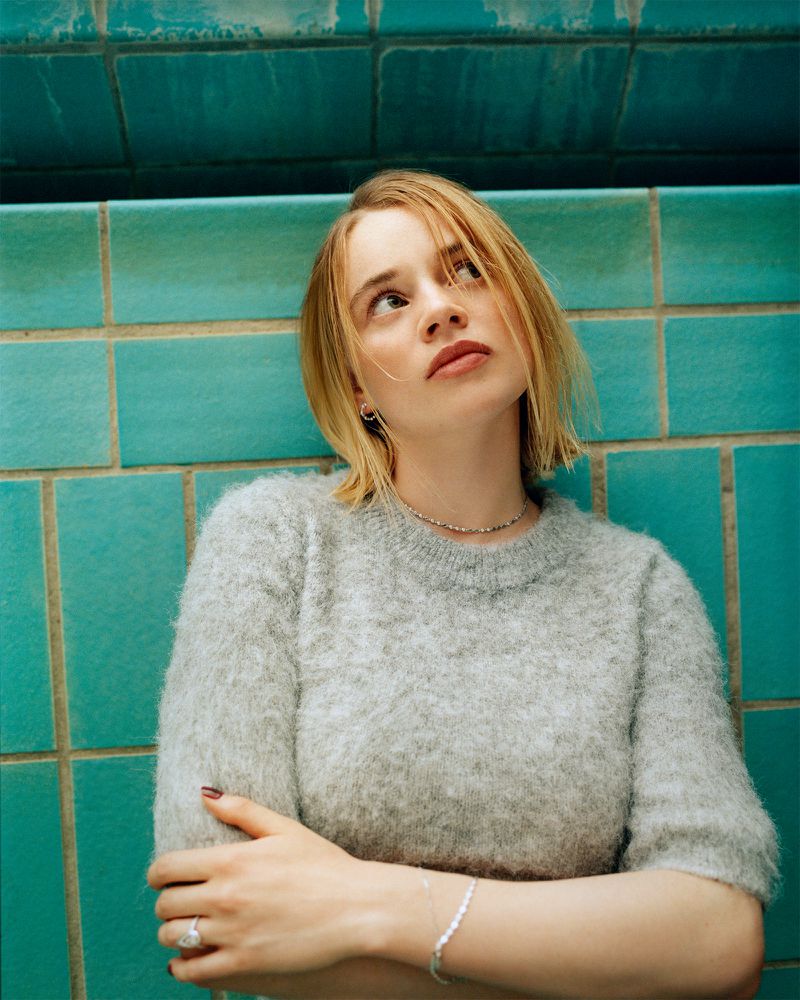 Close-up portrait of a young adult woman with blonde hair wearing a gray sweater, gazing upward next to a blue-tiled wall, beauty shot.