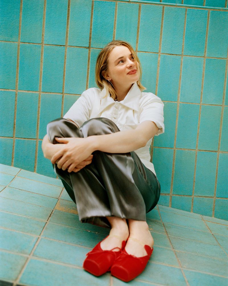 Luna Wedler seated on turquoise tiled steps wearing a white collared shirt, grey satin trousers, and red ballet flats.