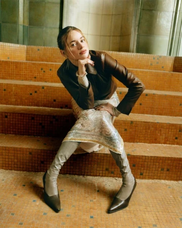 Luna Wedler seated on tiled terracotta stairs wearing a brown leather jacket, patterned skirt, and pointed boots for Vogue Germany.