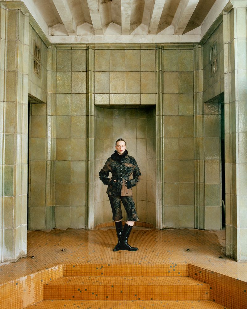A woman in a patterned jacket and shorts poses in a derelict tiled building with a neutral expression, lit by soft, natural light.