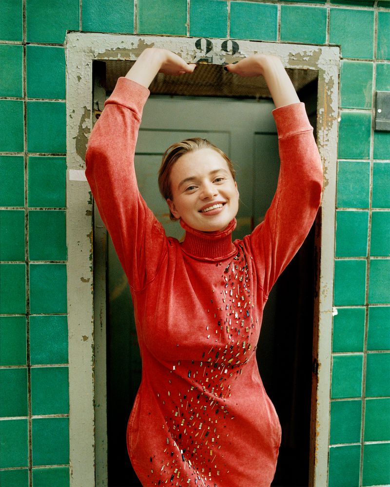 Woman in a red dress poses with arms raised in a doorway with green tile and the number 99 above, smiling and looking at the camera.