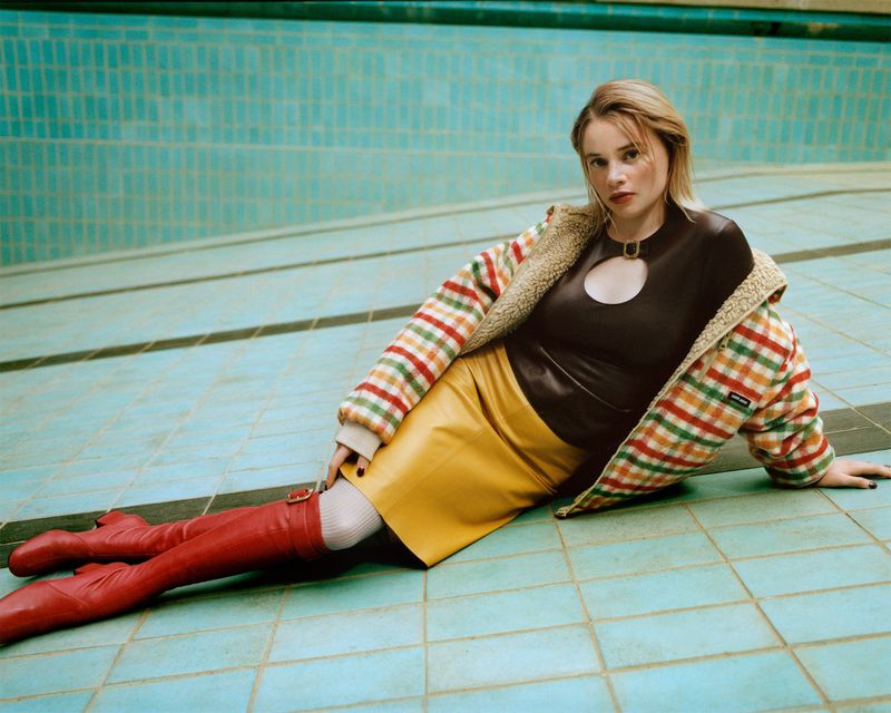 A fair-skinned woman with blonde hair poses in an empty swimming pool wearing a brown top, yellow skirt, and red boots.