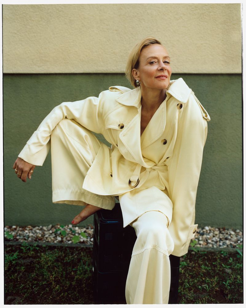 Adult woman with short blonde hair wearing a cream-colored pantsuit seated outdoors on a black suitcase with a serene expression.