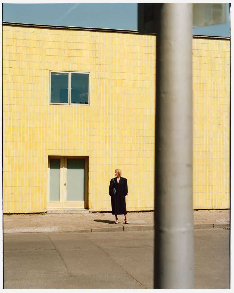 Full shot of a blonde woman in a black coat standing in front of a yellow building in an urban outdoor setting on a sunny day.