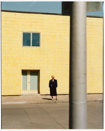 Full shot of a blonde woman in a black coat standing in front of a yellow building in an urban outdoor setting on a sunny day.