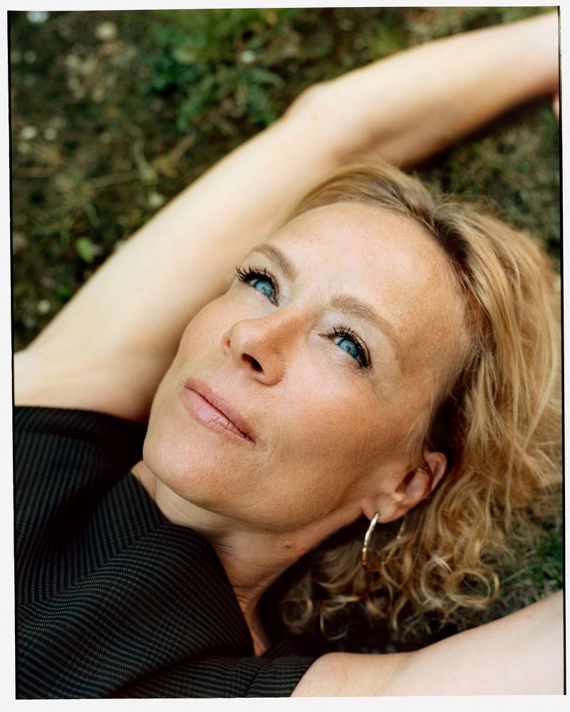 Close up of an adult woman with fair skin and blonde hair, lying on green grass and looking upwards with her arms resting behind her head.