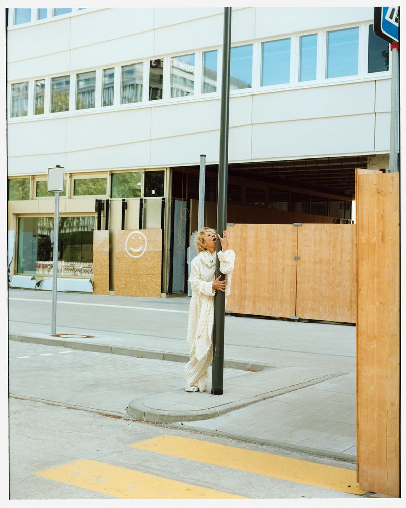 Adult woman in white dress stands on a street corner hugging a metal pole next to a pedestrian crosswalk in front of a light-colored building.