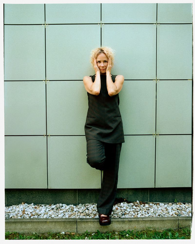 Woman in black pantsuit leans against a green tiled wall with hands on her face, outdoors on a sunny day, with small rocks visible.