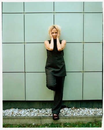 Woman in black pantsuit leans against a green tiled wall with hands on her face, outdoors on a sunny day, with small rocks visible.