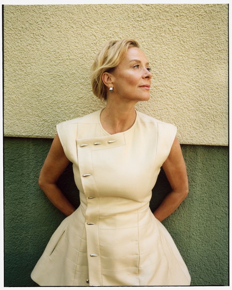 Portrait of an adult woman with short blonde hair, wearing an off-white dress, standing in front of a green and beige stucco wall.