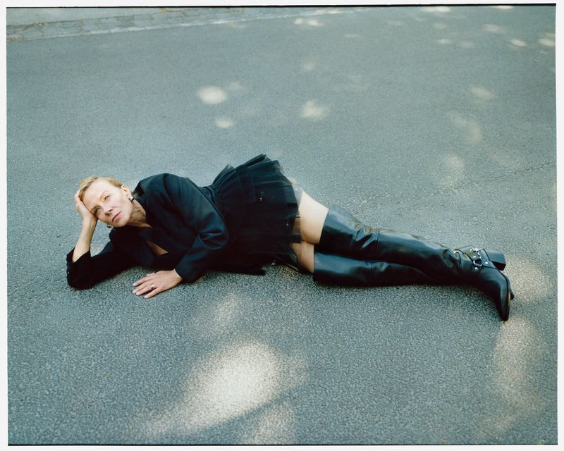 Adult woman in a black jacket and thigh-high boots lies on asphalt in the shade, striking a fashion pose with hand near her head.