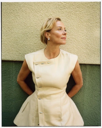 Katja Riemann wearing a structured cream sleeveless top with silver hardware against a textured two-tone stucco wall.