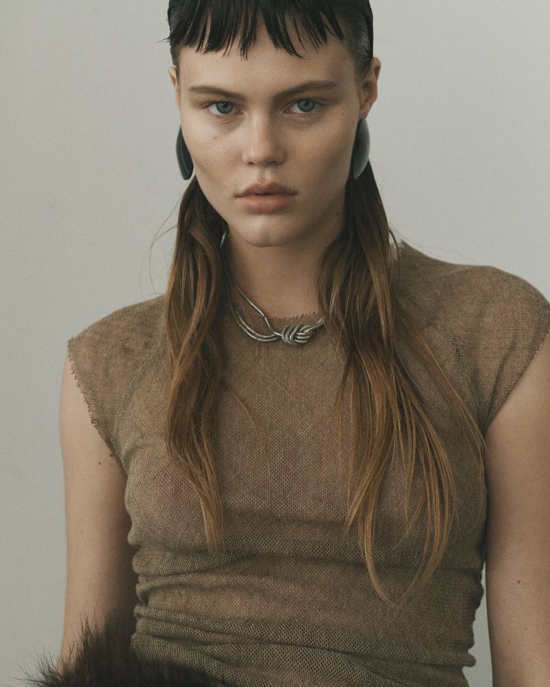 Close-up portrait of a female model in a textured brown dress, silver necklace, and dark earrings against a neutral background.