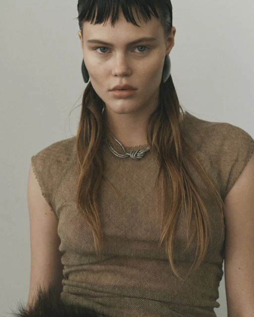 Close-up portrait of a female model in a textured brown dress, silver necklace, and dark earrings against a neutral background.